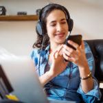 Happy young woman wearing headphones selecting a music playlist while working on laptop. Female using phone while working on laptop computer.